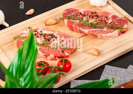 steaks de porc frais hachés avec épices et thym sur une planche à découper, à côté de tomates rouges mûres et d'ail Banque D'Images