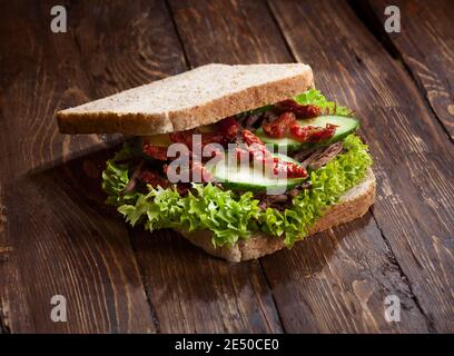 gros plan de sandwich au bœuf avec feuilles de laitue verte, tomates sèches, concombres frais et pain de seigle ou de blé, sur fond de table rustique foncé en bois, nobo Banque D'Images