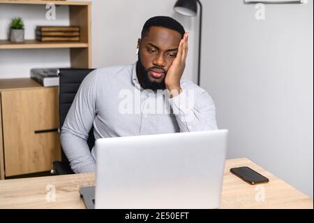 Jeune homme afro-américain se sentant stressé, fatigué et surtravaillé, tenant la tête dans les mains, assis à la réception devant un ordinateur portable, appliquant Banque D'Images