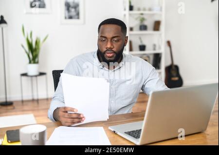 Un jeune propriétaire d'entreprise afro-américain concentré lisant attentivement le contrat, assis à la table et tenant des documents, vérifiant l'accord avec Banque D'Images