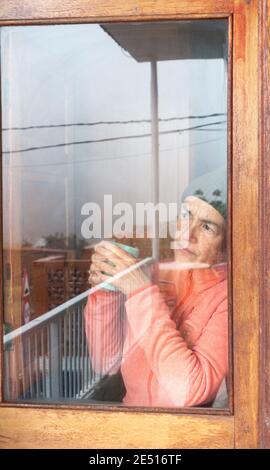 Femme d'âge moyen avec une tasse de café regardant par la fenêtre un jour froid. Banque D'Images