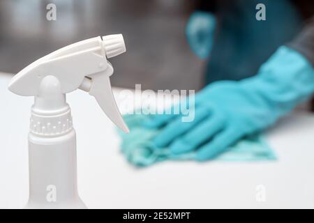 Flacon avec spray de désinfection au premier plan, personne floue avec des gants bleus à l'arrière pour nettoyer la surface blanche, pour prévenir l'infection par le covid-19 Banque D'Images