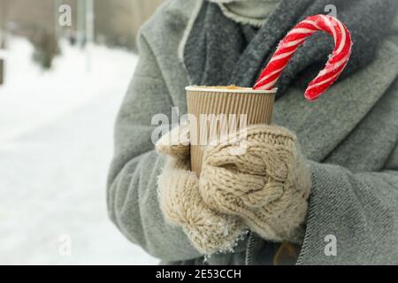 Femme en moufles tricotées tenant une tasse en papier de boisson chaude avec une canne en sucre à l'extérieur en hiver Banque D'Images