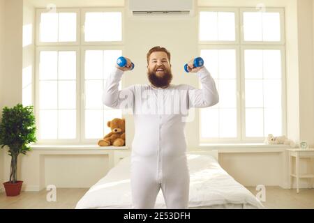 Joyeux sourire gras jeune homme en pyjama faisant de l'exercice de forme physique avec haltères à la maison Banque D'Images