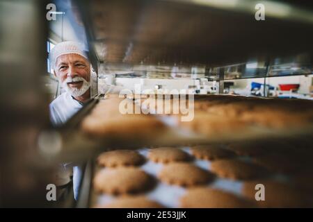 Photo d'homme boulanger adulte professionnel en uniforme de travail blanc. Debout devant les étagères avec des petits gâteaux frais. Banque D'Images