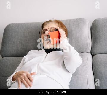 Une femme de 90 ans a été ravie de se détendre sur un canapé la pièce et parler sur un smartphone tout en regardant loin Banque D'Images