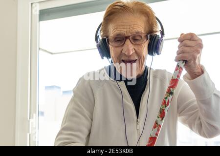 Femme âgée optimiste dans un casque d'écoute, écoutant de la musique et appréciant chansons tout en faisant des travaux ménagers avec un balai Banque D'Images