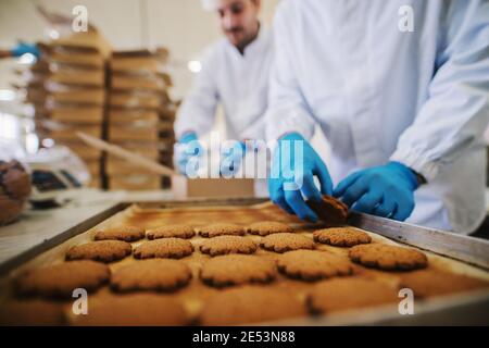 Gros plan sur le plateau rempli de petits gâteaux frais dans l'usine alimentaire. Image floue de deux hommes employés dans des vêtements stériles emballant des biscuits en arrière-plan. Banque D'Images