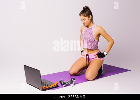 Une jeune fille sportive est assise sur le sol avec des haltères et utilise un ordinateur portable. Concept de formation en ligne. Arrière-plan blanc. Espace vide. Banque D'Images