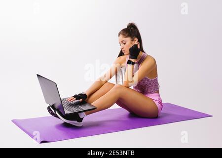 La femme sportive est assise sur un tapis de yoga avec un ordinateur portable. Concept de sport en ligne sur fond blanc. Banque D'Images