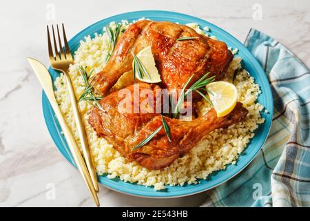 Des quartiers de cuisses de poulet servis sur le millet sur une assiette avec des couverts dorés, des quartiers de citron et des brins de romarin frais sur un fond de pierre en marbre blanc Banque D'Images