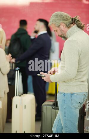 Homme mature debout avec des bagages dans une file d'attente et utilisant son téléphone portable en attendant le départ Banque D'Images