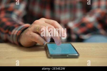 Un homme lit des nouvelles sur son téléphone portable, qui se trouve sur une table en bois. Gros plan sur l'anneau doré sur le doigt droit d'un homme Banque D'Images