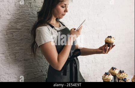 Une jeune blogueuse prend des photos sur son téléphone de délicieux gâteaux et cupcakes pour ses abonnés. Banque D'Images