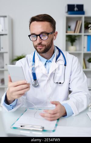Jeune médecin sérieux en whitecoat, écouteurs et lunettes tenant smartphone devant lui tout en consultant le patient en ligne Banque D'Images