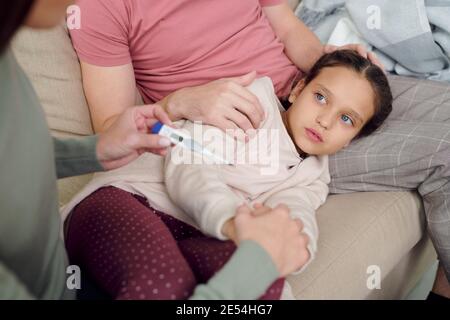 Main d'un jeune homme prudent prenant soin de malade adorable fille couchée sur un canapé à genoux pendant que sa femme mesure de la température du corps Banque D'Images