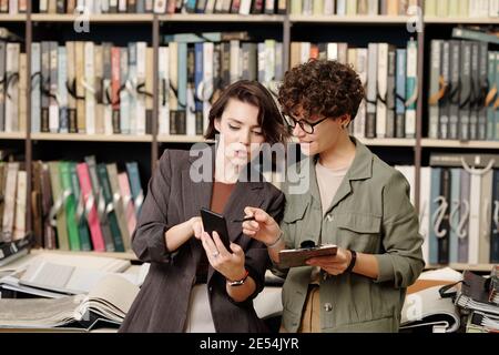 Jeune assistant de boutique souriant ou consultant de design studio pointage sur l'écran du smartphone tenu par le client féminin défilant échantillons Banque D'Images