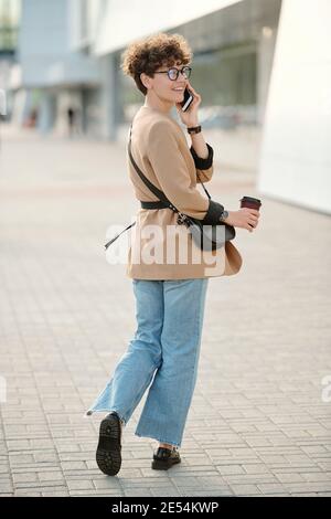 Vue arrière d'une jeune femme d'affaires contemporaine avec boisson et sac à main tenez votre smartphone à l'oreille tout en tournant et en regardant vers l'avant Banque D'Images