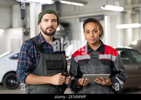 Deux jeunes travailleurs interculturels sérieux du centre de maintenance automobile debout devant l'appareil photo alors que l'un d'entre eux utilise une tablette en atelier Banque D'Images