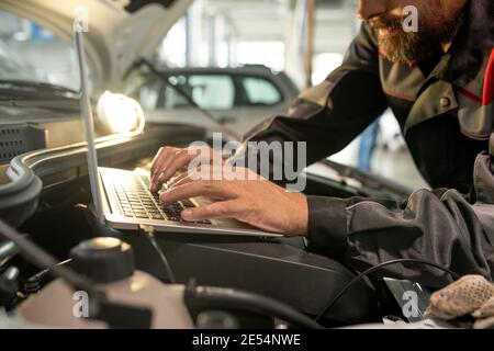 Mains d'un homme d'âge moyen du service d'entretien de voiture se pencher sur le compartiment moteur ouvert dans l'atelier tout en utilisant un ordinateur portable Banque D'Images