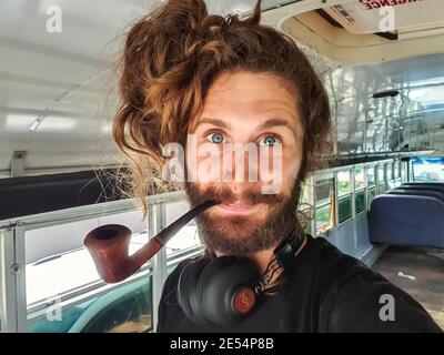 Portrait en gros plan d'un jeune homme curieux aux cheveux longs, avec de grands écouteurs et un tuyau vintage en bois à l'intérieur d'un bus en cours de rénovation Banque D'Images