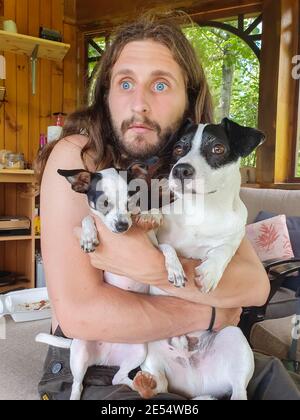 Jeune homme aux grands yeux bleus et aux longs cheveux blonds tenant serré à sa poitrine deux petits chiens noirs et bruns, un peu chihuahua et un terrier. Banque D'Images