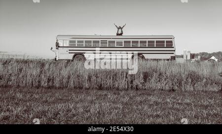 Photo grand angle d'un homme se tenant au sommet d'un bus au milieu de la campagne et levant ses bras au ciel. Noir et blanc. Banque D'Images