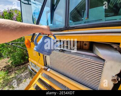 Gros plan de l'eau courante d'un homme avec un arrosoir bleu dans le radiateur d'un grand bus jaune. Entretien du véhicule. Banque D'Images