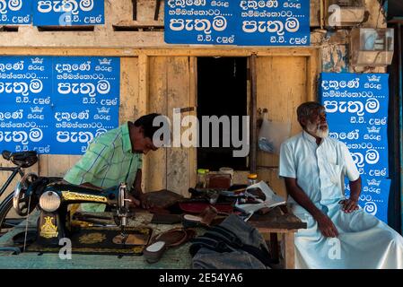 Tangalle, Sri Lanka : un cordonnier s'assoit et travaille en face de son magasin avec un ami Banque D'Images