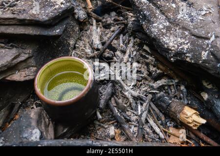 Gros plan de dessus d'une petite tasse de thé de chine verte avec de l'eau dans elle reposant sur les cendres maintenant froides de ce qui était un petit feu de camp. Banque D'Images
