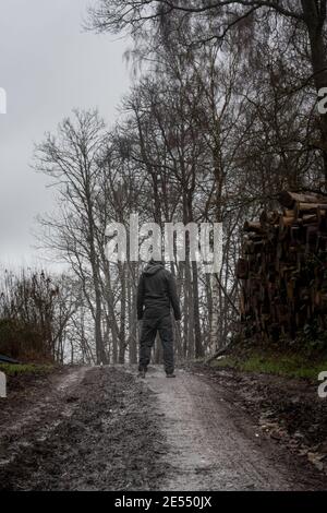 Un homme à capuchon, de retour à la caméra sur un sentier boueux, à côté d'une pile de bois sur un gris moody jour dans la campagne Banque D'Images