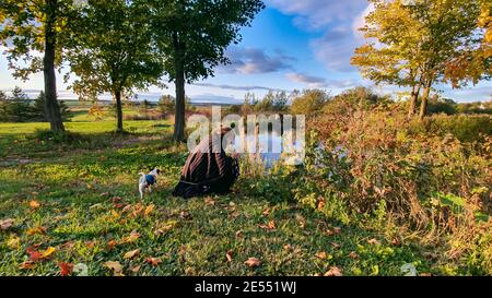 Photo grand angle d'une jeune femme avec son petit chien agenouillé pour examiner quelques arbustes qui poussent sur le bord d'un petit étang dans la campagne. Banque D'Images
