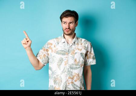 Mécontent touriste dans une chemise hawaïenne, regardant et pointant à gauche déçu, se plaignant quelque chose de mauvais, debout sur fond bleu Banque D'Images