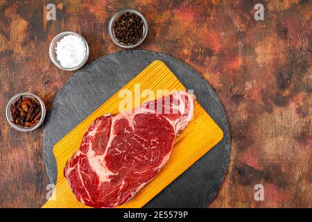 Sur une plaque en ardoise ronde, le bœuf cru est agrémenté de récipients en verre contenant des grains de poivre, du sel grossier et des piments cayenne sur une ancienne table en bois, en brun Banque D'Images