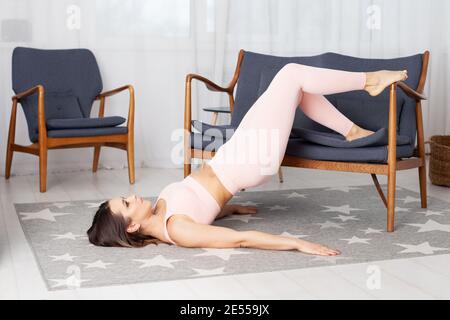 Une jeune femme pleine longueur en leggings de sport roses pose mentir sur le sol et en soulevant ses jambes sur un gris intérieur de la maison avec canapé rembourré Banque D'Images