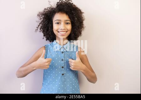 Une jeune fille de pretteen biracial joyeuse montre des pouces vers le haut, une écolière portant une chemise à pois regarde l'appareil photo et les sourires, isolée sur rose pastel. Réussite Banque D'Images