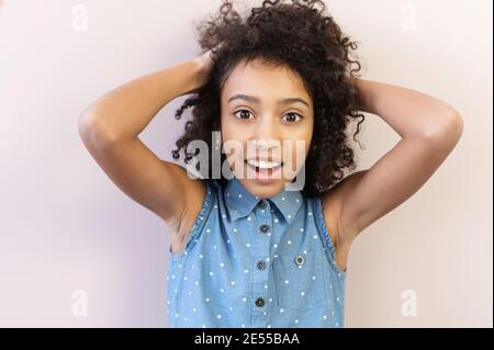 Portrait en gros plan d'une jolie jeune fille africaine surprise, une écolière regarde l'appareil photo avec les yeux grands ouverts et les cheveux avec les mains Banque D'Images