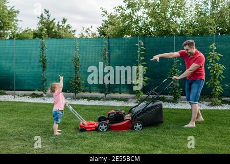 Le père et son fils s'amusent à tondre la pelouse ensemble. Longueur complète d'un jeune garçon heureux coupant l'herbe avec son père. Banque D'Images