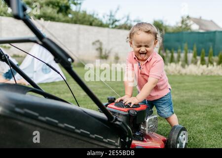 Portrait d'un jeune enfant avec ses yeux fermés et la langue sortie debout à une tondeuse à gazon. Un tout-petit qui s'amuse à couper de l'herbe. Banque D'Images