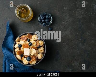 Plats tendance - crêpes, céréales, beurre et bleuets servis. Tas de mini crêpes aux céréales dans le boul sur fond sombre. Copier l'espace vers la droite pour le texte. Vue de dessus ou plan de travail. Banque D'Images