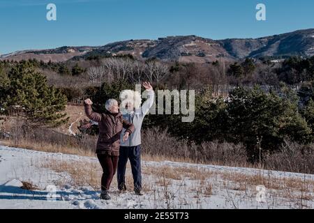 Heureux couple d'âge mûr dansant dans la nature sur fond de montagnes. Une femme aux cheveux gris dans une veste et un pantalon marron chauds. Un homme dans un traditi Banque D'Images
