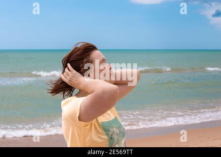 une femme adulte à la retraite se tient sur la mer tournée pour faire face au vent et redresse ses cheveux, bonne heure d'été sur la côte, la fin de l'auto-isolement et de la quarantaine Banque D'Images