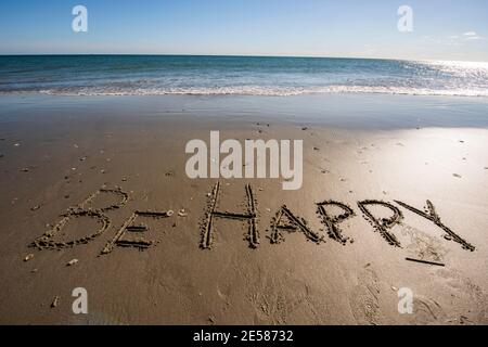 Les mots ÊTRE HEUREUX écrit dans le sable au bord de la mer. Banque D'Images