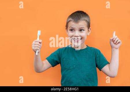 Un garçon sans la dent supérieure de lait dans un t-shirt vert tient la brosse à dents à la main sur fond orange. Banque D'Images