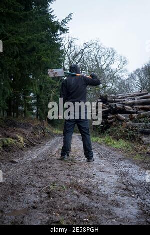 Un homme à capuchon, de retour à la caméra sur un sentier boueux tenant une bêche, à côté d'une pile de rondins lors d'un jour gris moody dans la campagne Banque D'Images