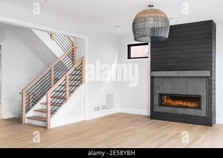 Un beau salon neuf et vide avec un lustre lumineux en forme de panier suspendu devant une cheminée gris foncé. Un escalier personnalisé possède des broches en fer. Banque D'Images