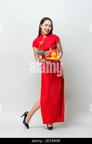 Jeune femme asiatique en robe de cheongsam rouge traditionnel tenant la chance Fruits pour la nouvelle année chinoise dans un studio isolé gris clair arrière-plan Banque D'Images