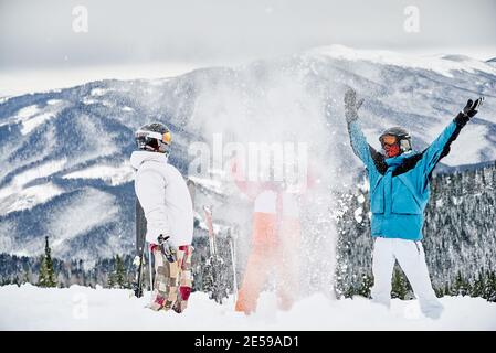 Amis skieurs en costume de ski jetant de la neige poudreuse fraîche en plein air. Deux hommes et une femme s'amusent sur une colline enneigée avec de belles montagnes en arrière-plan. Concept de station de ski et d'amitié. Banque D'Images