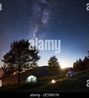 Camping touristique près de la forêt la nuit. Deux tentes lumineuses et feu de joie sous un ciel nocturne incroyable plein d'étoiles et de voie lactée. Sur les arbres de fond, beau ciel étoilé, montagnes et pleine lune Banque D'Images