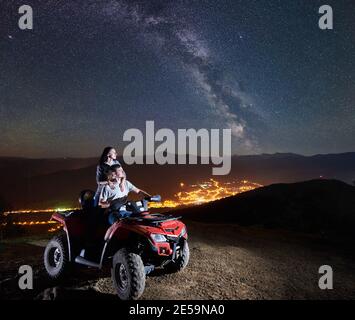 Jeune couple homme et femme touristes à cheval VTT quad moto ensemble sur le sommet de la montagne, appréciant la vue magnifique du ciel de nuit plein d'étoiles, voie lactée, ville lumineuse sur fond Banque D'Images